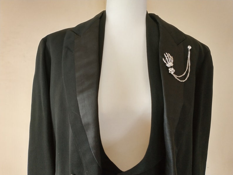 May include: A black tuxedo jacket with a silver skeleton hand lapel pin. The pin has a chain attached to it.