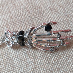 May include: A silver skeleton hand pendant necklace with black rhinestone accents. The necklace has a chain and a decorative clasp.