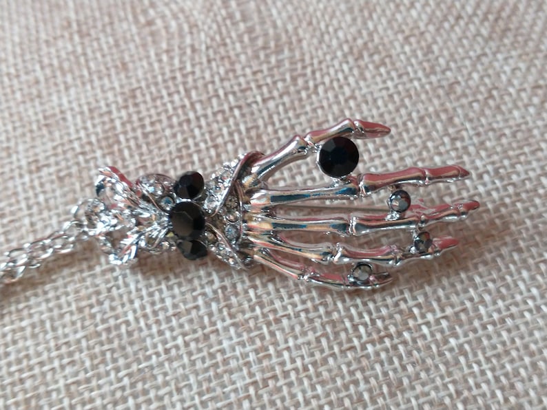 May include: A silver skeleton hand pendant necklace with black rhinestone accents. The necklace has a chain and a decorative clasp.