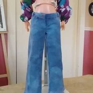 Pode incluir: Uma boneca vestindo um top cropped com estampa floral com mangas compridas e calças boca de sino de ganga azul. As calças têm bainha desfiada. A boneca está em um suporte branco.