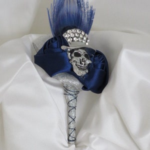 May include: A silver skull with rhinestones and a blue feather is attached to a bouquet of blue satin roses. The bouquet is wrapped in silver glitter and has a blue ribbon.
