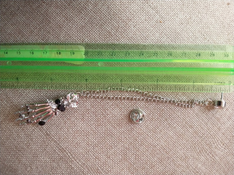 May include: A silver skeleton hand charm with black rhinestones attached to a silver chain with a safety pin clasp.