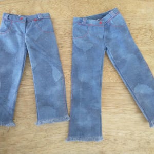 May include: Two pairs of light blue denim jeans with red stitching and frayed hems. The jeans have a faded wash effect and classic five-pocket styling. The jeans are displayed on a wooden surface.