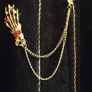 May include: A gold skeleton hand pin with red rhinestone accents attached to a gold chain.