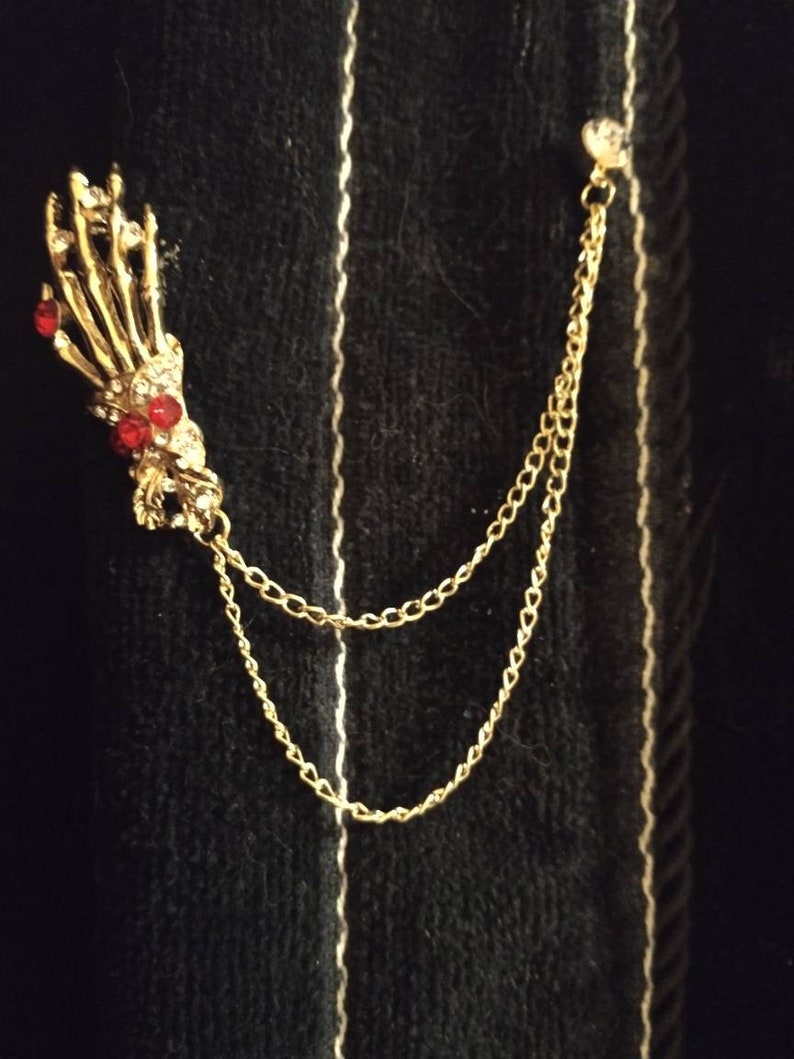 May include: A gold skeleton hand pin with red rhinestone accents attached to a gold chain.