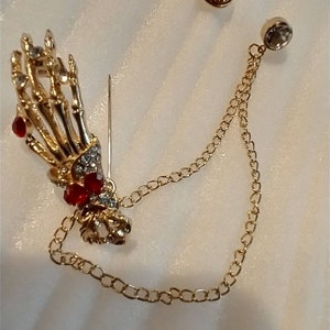 May include: A gold skeleton hand brooch with red and clear rhinestones, attached to a gold chain with a clear rhinestone at the end.