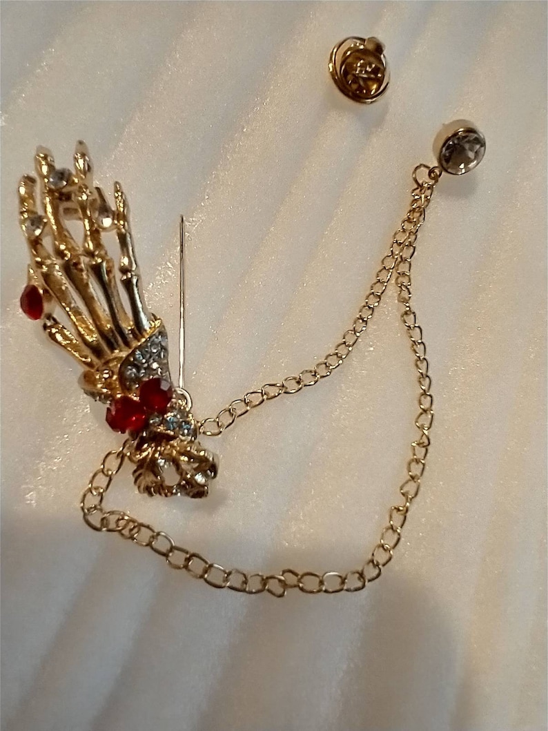 May include: A gold skeleton hand brooch with red and clear rhinestones, attached to a gold chain with a clear rhinestone at the end.