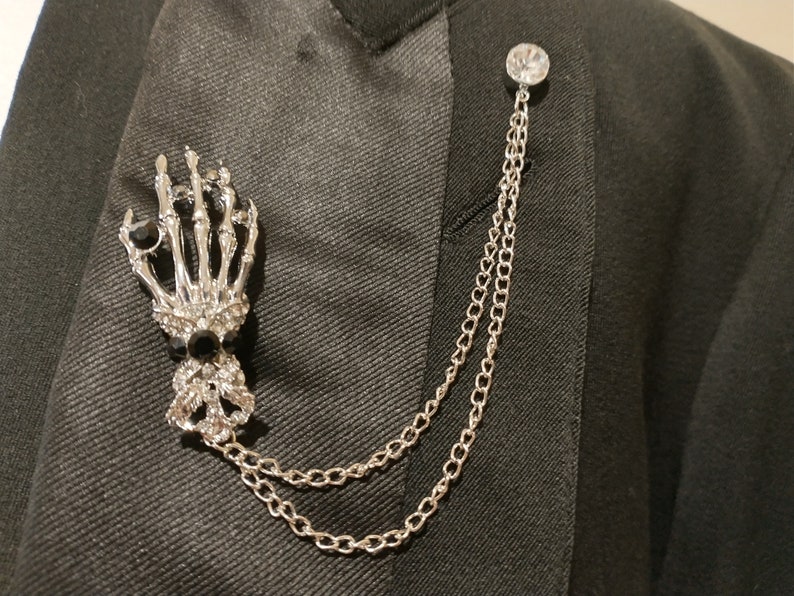 May include: A silver skeleton hand lapel pin with a clear rhinestone on the top of the hand. The pin is attached to a silver chain with a clear rhinestone at the top.