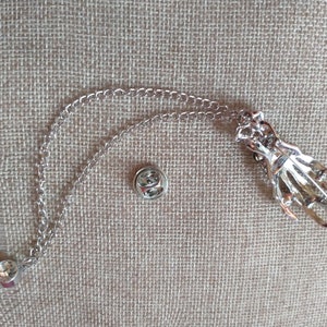 May include: Silver skeleton hand pin with a chain attached. The pin has a safety clasp and the chain has a loop at the end.