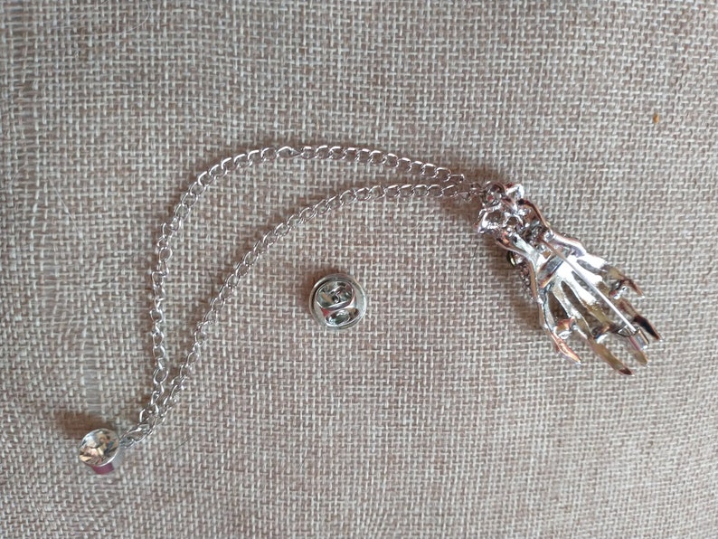 May include: Silver skeleton hand pin with a chain attached. The pin has a safety clasp and the chain has a loop at the end.