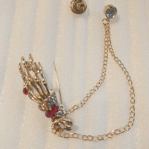 May include: A gold skeleton hand brooch with red and clear rhinestones, attached to a gold chain with a clear rhinestone at the end.