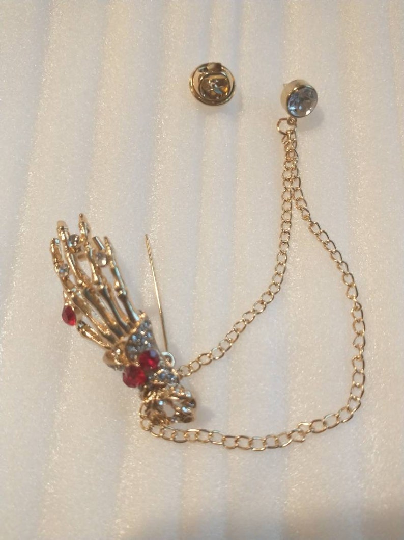 May include: A gold skeleton hand brooch with red and clear rhinestones, attached to a gold chain with a clear rhinestone at the end.