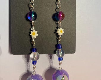 May include: A pair of dangle earrings with silver-tone hooks. Each earring features iridescent beads, small white daisy charms, clear and blue beads, and a purple ball with the number 4. The earrings are suspended from a silver chain.