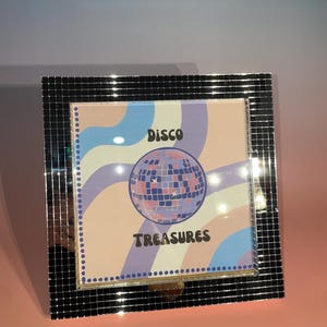 May include: A square, mirrored picture frame with a colourful print. The print features a disco ball graphic and the words "Disco Treasures" in black. The background has pastel colours of pink, blue, and purple.