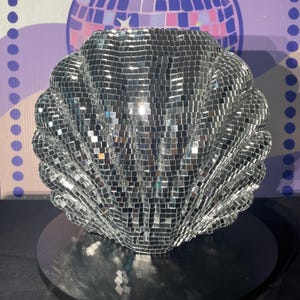 May include: A decorative, silver disco ball seashell-shaped object. The shell is covered in small, reflective squares, creating a shimmering effect. It sits on a black, circular base, with a disco ball graphic in the background.