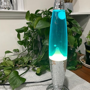 Könnte beinhalten: Eine blaue Lavalampe mit einem silbernen Mosaikfuß und einem silbernen Mosaikdeckel. Die Lampe steht auf einer weiß-grauen, gemusterten Oberfläche.