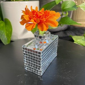 May include: A small, rectangular vase covered in mirrored tiles, holding a vibrant orange flower. The vase has a short, cylindrical neck. The flower is a bright, multi-petaled bloom. The background includes green plants.