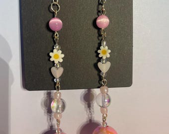 May include: A pair of dangle earrings with silver hooks. Each earring features a pink cat-eye bead, a white daisy, a clear heart, clear beads, and a pink ball with the number "11" and "1" printed on them.