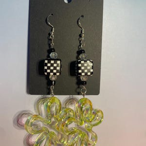 May include: A pair of dangle earrings with a black and white checkered square bead, clear and black beads, and a large, iridescent, flower-shaped charm. The earrings hang from silver-colored hooks.