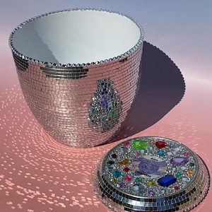 May include: A cylindrical container covered in small silver mirror tiles with a white interior. The matching circular lid features a border of mirror tiles and a center decorated with colorful faceted gems and a label reading Disco Treasures.