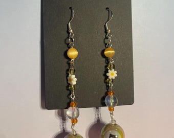 May include: Dangle earrings with a silver hook, featuring yellow beads, small white daisy-shaped beads, and clear beads with orange accents. The earrings end with a round, iridescent bead with the number 7.