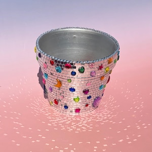 May include: A cylindrical planter covered in small square silver mirror tiles, creating a disco ball effect. The exterior is decorated with various colorful, faceted plastic gems in shapes including hearts, stars, and moons, with a silver rim.