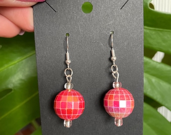 May include: A pair of dangle earrings featuring red and pink mosaic-style beads. The beads are spherical and have a faceted surface. The earrings hang from silver-colored hooks and are displayed on a black card.
