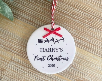 Puede incluir: Adorno navideño de cerámica blanca con una cinta roja y una silueta negra del trineo de Papá Noel con renos. El adorno dice "HARRY'S First Christmas 2020".