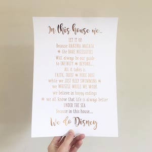 May include: A white paper print with rose gold text that reads "In this house we..." followed by Disney-themed quotes. The print is held by a hand and is against a neutral background. The text includes phrases like "Let it go" and "We do Disney."