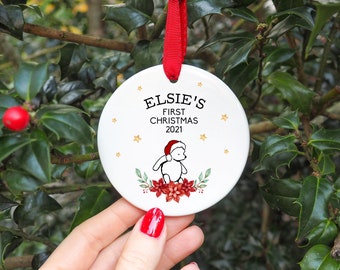 Op de afbeelding: Witte kerstbal van keramiek met een rood lint. De kerstbal heeft een zwart-witte illustratie van een beer met een kerstmanmuts, omringd door rode kerststerren en groen blad. De tekst "ELSIE'S FIRST CHRISTMAS 2021" is in zwart op de kerstbal gedrukt.