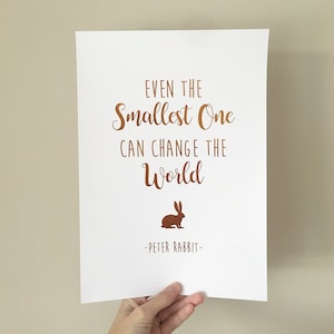 Puede incluir: Un póster blanco con una cita en color cobre que dice "Incluso el más pequeño puede cambiar el mundo - Peter Rabbit" con una silueta de conejo en color cobre.