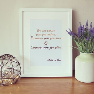 Op de afbeelding: Een wit ingelijst print met een gouden en bruine inspirerende quote van Winnie de Poeh. De quote luidt "Je bent dapperder dan je denkt, sterker dan je lijkt en slimmer dan je denkt".