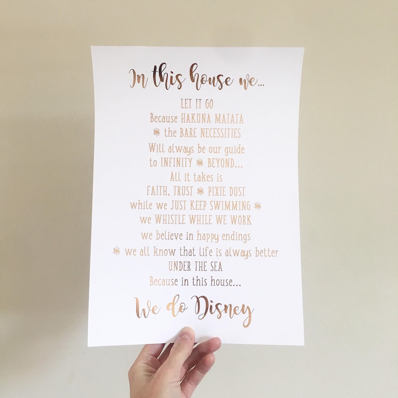 May include: A white paper print with rose gold text that reads "In this house we..." followed by Disney-themed quotes. The print is held by a hand and is against a neutral background. The text includes phrases like "Let it go" and "We do Disney."