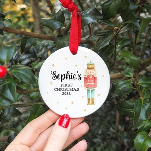 May include: White ceramic Christmas bauble with a gold star pattern. It features a cartoon illustration of a nutcracker and the text "Sophie's First Christmas 2022".