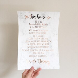 May include: A white paper print with a marble-like background and rose gold text. The text reads "In this house we..." followed by quotes from Disney movies. The bottom of the print says "We do Disney".