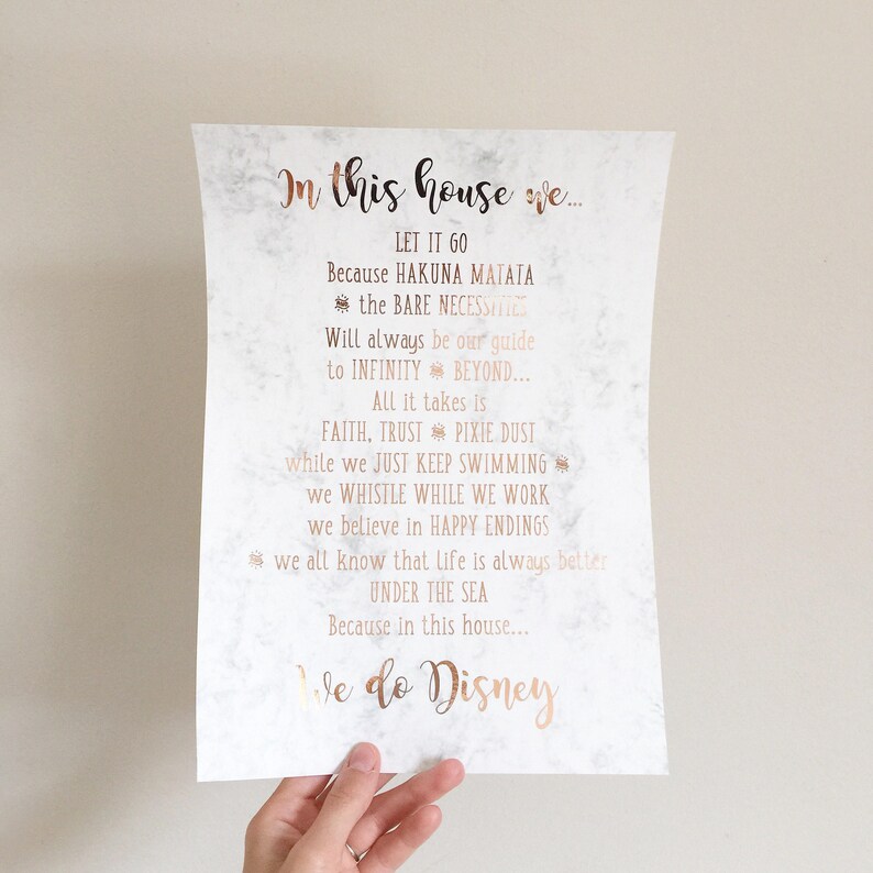 May include: A white paper print with a marble-like background and rose gold text. The text reads "In this house we..." followed by quotes from Disney movies. The bottom of the print says "We do Disney".