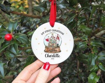 May include: White ceramic Christmas bauble with a gold snowflake design. The bauble features a snow globe with a ballerina, a nutcracker, and a Christmas tree inside. The text "My First Christmas" and "Charlotte 2022" is printed on the bauble.