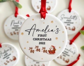 Puede incluir: Adorno navideño de cerámica blanca con una cinta roja. El adorno presenta a un Papá Noel en un trineo tirado por renos, estrellas doradas y el texto "Amelia's First Christmas 2022".