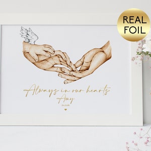 May include: A white framed print with a watercolour illustration of two hands holding each other. The hands are brown and the print has the text "Always in our hearts Amy 21.11.20" in gold foil.