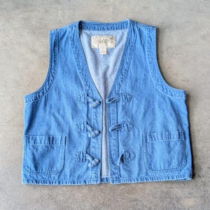 May include: Light blue denim vest with a V-neck, two front pockets, and toggle closures. The vest has a light wash and a vintage aesthetic. The vest is sleeveless and has a label on the inside.