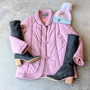 May include: A pink quilted jacket with a button closure and a high collar. The jacket is paired with black knee-high boots and a light blue and pink knit beanie with a fluffy pom-pom.