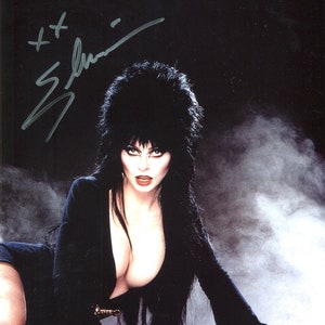 May include: A woman with long black hair and dark makeup is wearing a black dress. She is posing on a white surface with a smoky background. The image is signed "Elvira".