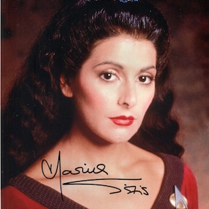 May include: A portrait of a woman with dark hair and red lipstick, wearing a red and brown top. The image includes an autograph and the Star Trek: The Next Generation logo.