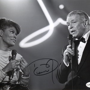 Peut inclure: Photographie en noir et blanc de deux personnes, un homme et une femme, tenant chacune un microphone. L'homme porte un smoking et un nœud papillon. La femme porte un haut à manches longues. L'image est autographiée.
