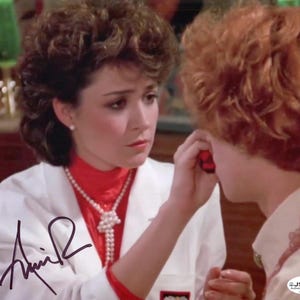 May include: A signed photograph featuring two women. One woman, wearing a white lab coat over a red turtleneck and pearl necklace, is looking at the other woman. The second woman has red hair and is wearing a light-colored shirt.