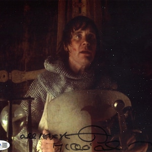 Peut inclure: Photographie signée d'un homme en cotte de mailles, tenant un bouclier. L'image a une esthétique vintage, avec des signes d'usure. La signature et l'inscription "all love" sont présentes. Un autocollant d'authentification JSA est visible.