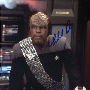 May include: A color photograph of a man in a Star Trek uniform. He has a distinctive forehead, a beard, and is wearing a sash with a silver chain design. The image includes an autograph in blue ink.