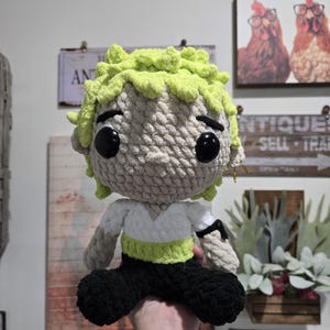 Op de afbeelding: Handgemaakte gehaakte pop met lichtgroen haar, grote zwarte ogen en een neutrale huidskleur. De pop draagt een wit shirt met groene en zwarte bies en een zwarte broek. Het is een handgemaakt item.