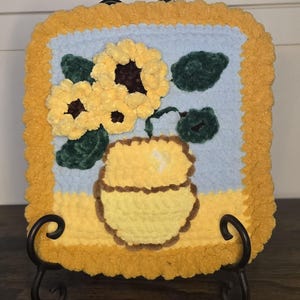 Könnte beinhalten: Ein handgefertigtes, quadratisches Häkelstück mit einem Sonnenblumen-Design. Die Sonnenblumen sind gelb mit braunen Zentren, vor einem hellblauen Hintergrund. Das Stück ist mit einem gelben Rand eingefasst und auf einem schwarzen Metallständer ausgestellt.