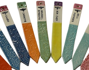May include: Set of ten colourful ceramic garden markers with bee and flower designs. Each marker has a different vegetable name written on it:  'Chard', 'Carrots', 'Brussel Sprouts', 'Bok Choy', 'Bell Peppers'.